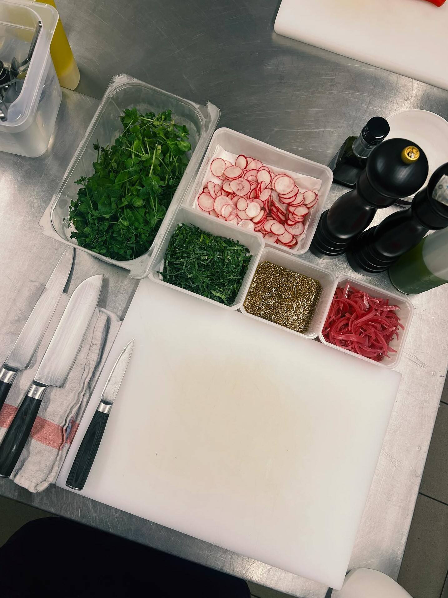 Mise en place en cuisine, vue du dessus : jeunes pousses, radis en fines rondelles, herbes ciselées, graines et lamelles d’oignon rouge en bacs, à côté des moulins à poivre et des couteaux.