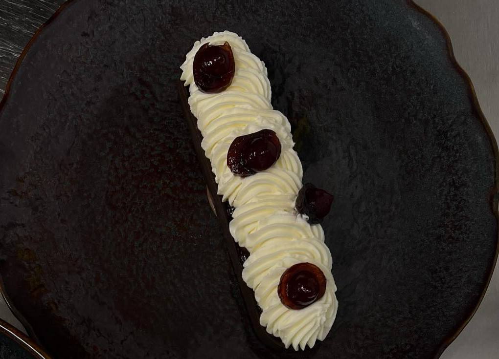 Forêt-noire en longueur, chantilly pochée et cerises amarena, sur une assiette sombre.