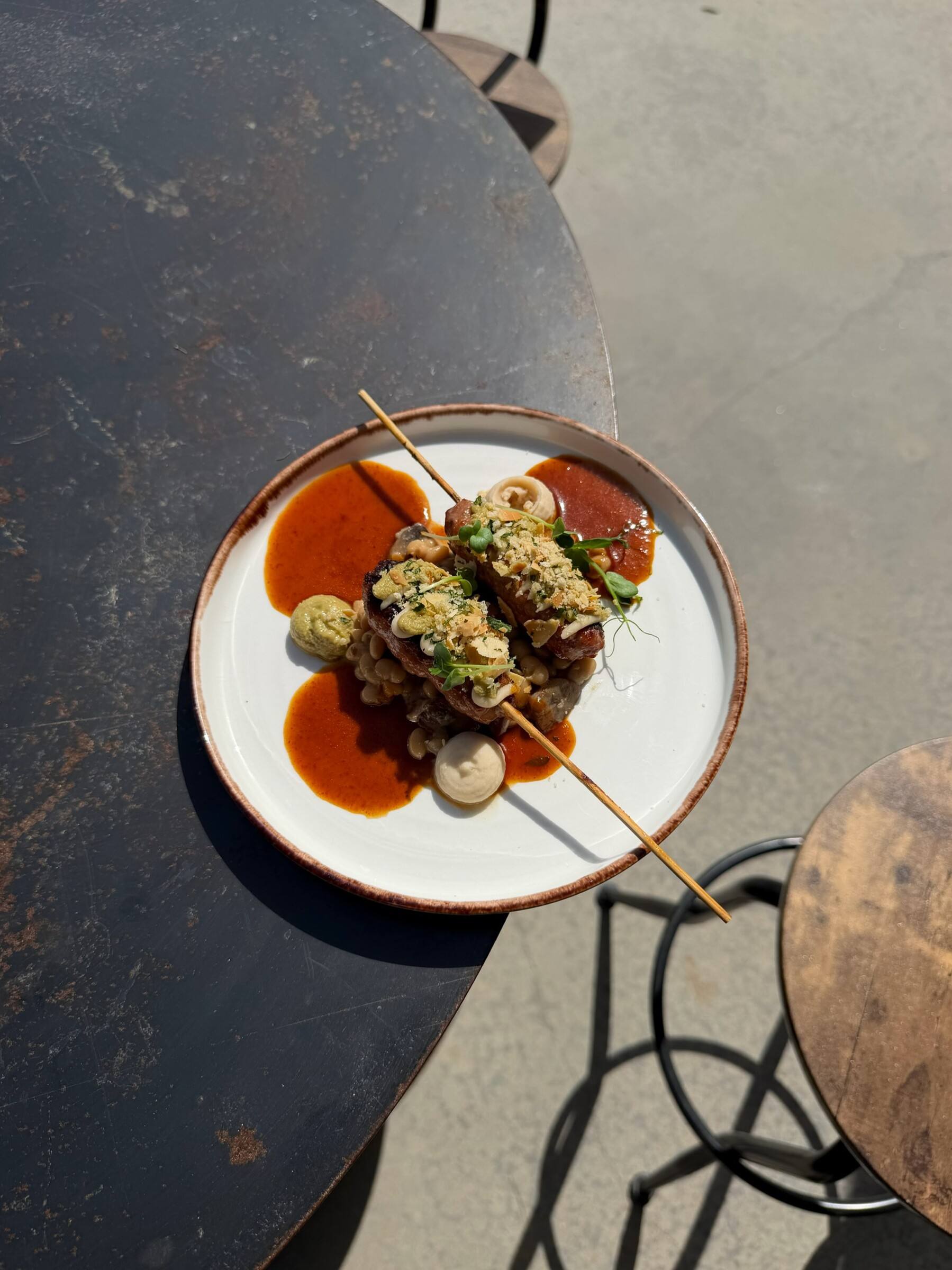 Brochette posée sur une assiette blanche, sauce rouge et condiment, sur une table en métal au soleil.