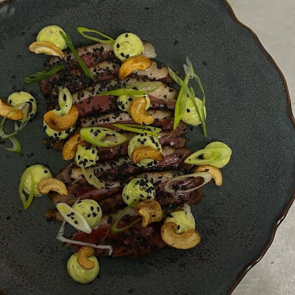 Tataki de magret vu du dessus : tranches de magret, noix de cajou et points de mayonnaise à l’estragon, sur une assiette sombre.
