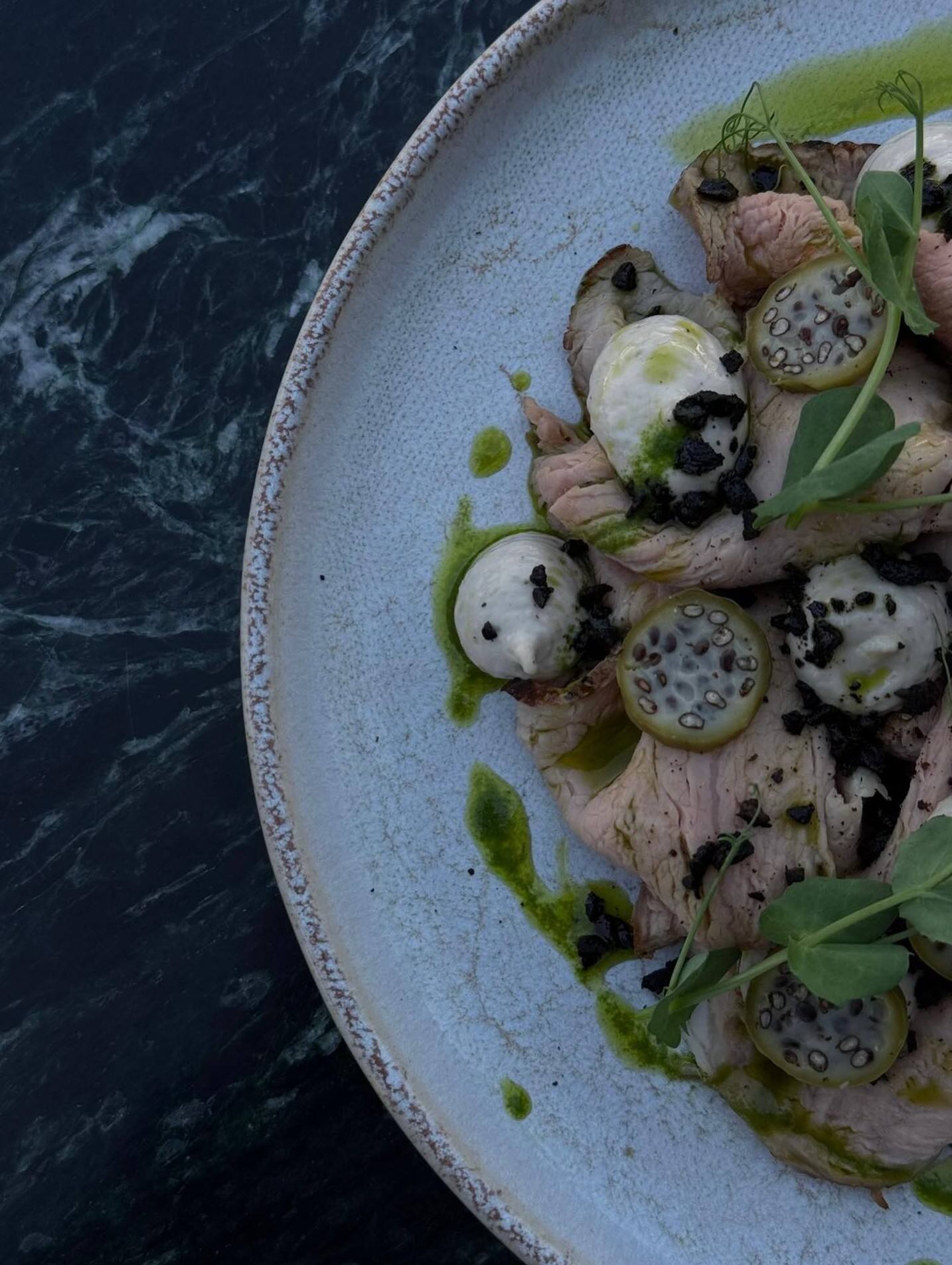 Détail du vitello tonnato dressé, vu du dessus : veau tranché en vagues, points de sauce, câpres à queue en rondelles, poudre d’olive, huile de roquette et pousses, sur une assiette en grès claire.