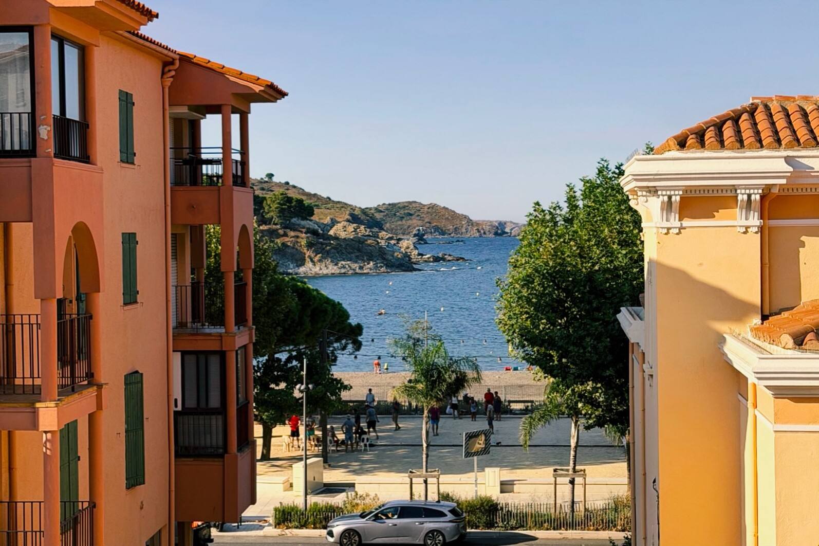 Un village au bord de la mer en plein soleil : façades ocre, une place devant la plage, la baie et ses rochers au fond.